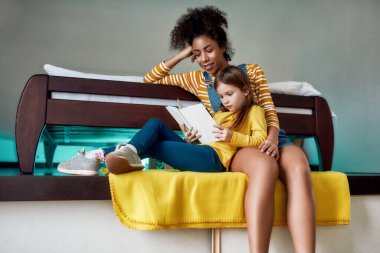 Anlar. Afro-Amerikan kadın, bebek bakıcısı ve kafkasyalı şirin küçük kız evde otururken birlikte masal okuyorlar. Çocuk eğitimi, boş zaman aktiviteleri, bebek bakıcılığı konsepti