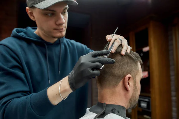 Peluquero trabajando en la peluquería. Joven peluquero con gorra haciendo corte de pelo para su cliente en la peluquería. Corte de pelo para hombre —  Fotos de Stock