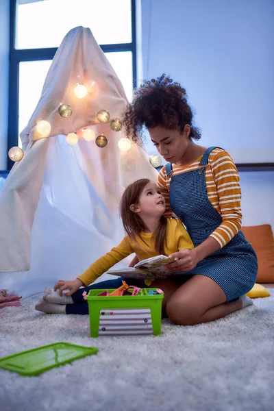 Una babysitter di cui ti puoi fidare. Donna afroamericana baby sitter divertente caucasico carina bambina. Bambino che legge un libro con la tata seduta in wigwam, tenda — Foto Stock