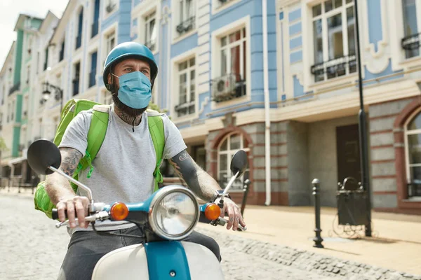 Bezahlbar. Liefermann mit Helm, der aufgrund des Covid19-Virus eine Maske trägt, auf einem Motorroller fährt und Essen ausliefert. Kurier, Lieferservice, Lockdown-Konzept — Stockfoto