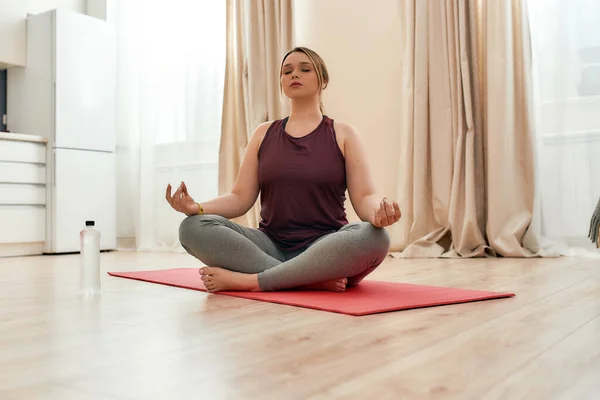 Zihnini rahatlat. Spor kıyafetleri içinde, evde yoga minderinde lotus pozisyonunda meditasyon yapan kıvrımlı bir kadın. — Stok fotoğraf