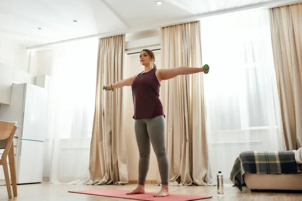 Ponte en forma. Tiro de cuerpo entero de mujer joven con curvas en ropa deportiva haciendo ejercicio usando pesas en una esterilla de yoga en casa — Foto de Stock