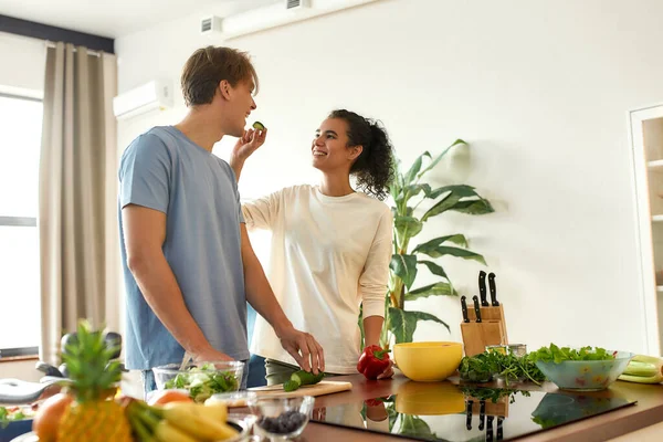 A fiatal férfi zöldségeket szeletel, míg a nő uborkával eteti. A vegetáriánusok egészséges ételt készítenek együtt a konyhában. Vegarianizmus, egészséges étel koncepció — Stock Fotó