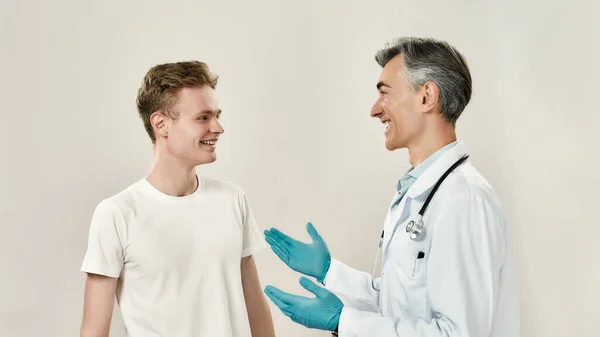 Všechno je v pořádku. Starší veselý mužský lékař v lékařské uniformě a modré sterilní rukavice mluví se šťastným mladým mužským pacientem. Diskusní diagnóza — Stock fotografie