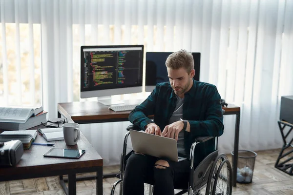 Multitasken. Portret van een jonge mannelijke webontwikkelaar in een rolstoel die programmacode schrijft op zijn laptop op zijn werkplek op kantoor. Meerdere computerschermen met programmacode — Stockfoto