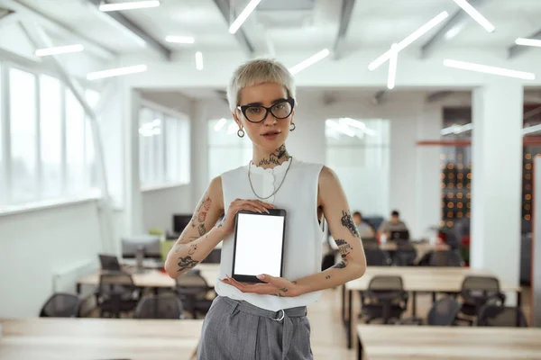 New gadget. Portrait of young and attractive blonde tattooed businesswoman in eyeglasses showing digital tablet and looking at camera while standing in modern working space