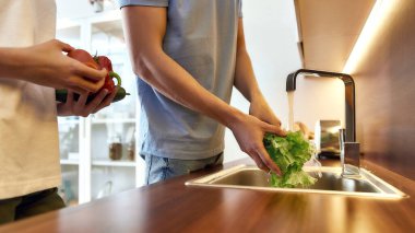 Yemek pişirmeden önce adamın elleri modern mutfak lavabosunda marul yıkıyor, yemek hazırlıyor. Kadın ona yardım ediyor, diğer sebzeleri tutuyor. Vejetaryenlik, sağlıklı gıda, hijyen konsepti