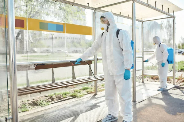 Virusbusters. Sanitization, cleaning and disinfection of the streets and alleys in the city due to the emergence of the Covid19 virus. Men in protective suits and masks at work near the bus stop — Stock Photo, Image