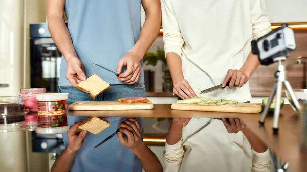 Μερικοί χορτοφάγοι καταγράφουν ένα video blog ενώ ετοιμάζουν υγιεινό γεύμα στην κουζίνα μαζί. Άντρας απλώνει χούμους πάνω σε φρυγανισμένο ψωμί ενώ η γυναίκα κόβει αβοκάντο — Φωτογραφία Αρχείου