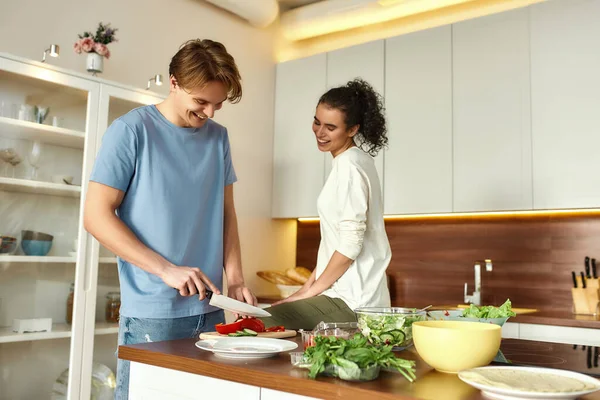 Νεαρός άντρας χαμογελάει, κόβει λαχανικά ενώ η γυναίκα μιλάει και τον παρακολουθεί. Χορτοφάγοι ετοιμάζουν υγιεινό γεύμα στην κουζίνα μαζί. Χορτοφαγία, υγιεινή διατροφή έννοια — Φωτογραφία Αρχείου
