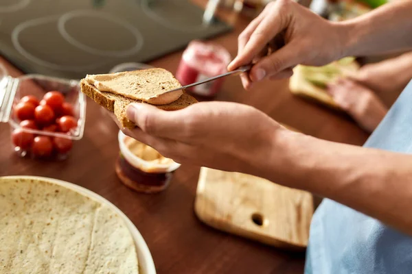 Κοντινό πλάνο του ανθρώπου που απλώνει χούμους πάνω σε φρυγανισμένο ψωμί. Μερικοί χορτοφάγοι ετοιμάζουν υγιεινό γεύμα στην κουζίνα μαζί. Χορτοφαγία, υγιεινή διατροφή έννοια — Φωτογραφία Αρχείου