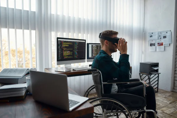 Joven desarrollador web masculino en silla de ruedas con gafas de realidad virtual mientras trabaja en su lugar de trabajo en la oficina moderna. Múltiples pantallas de ordenador con código de programa —  Fotos de Stock