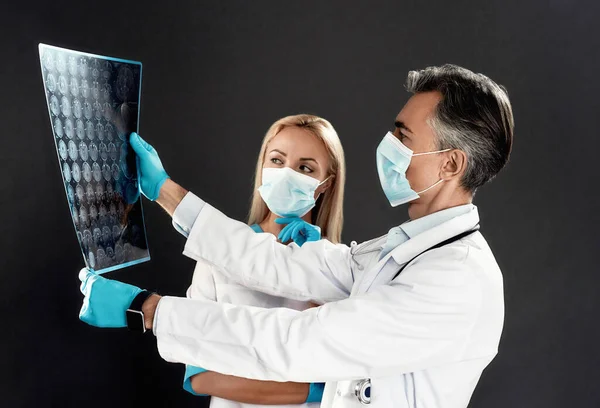Tıbbi üniformalı ve koruyucu maskeli kadın ve erkek doktorların röntgene bakması, CT taramasını inceleyip karanlık arka planda durmaları. — Stok fotoğraf