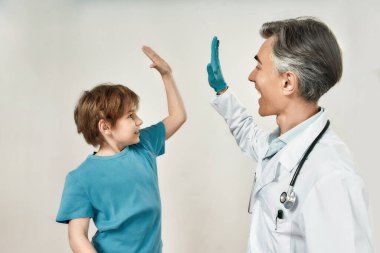 Başarılı bir aşıdan sonra. Tıbbi maskeli ve mavi steril eldivenli dost canlısı, olgun doktor çocuk doktoru mutlu küçük bir çocuğa beşlik çakıyor.