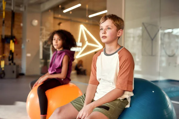 Teenage boy looking at trainer while sitting on exercise ball with other kids in gym. Sport, healthy lifestyle, physical education concept — Stock Photo, Image