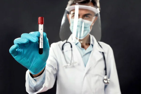 Μόλυνση από τον ιό Κορόνα. Γιατρός με ιατρική μάσκα και ασπίδα προσώπου που κρατάει δοκιμαστικό σωλήνα με αίμα για εξέταση COVID-19. Κύρια εστίαση σε δοκιμαστικό σωλήνα — Φωτογραφία Αρχείου