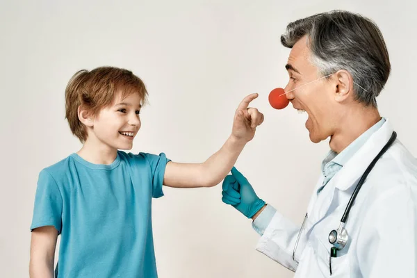 Emocionado menino tocando nariz de palhaço de pediatra masculino amigável, isolado sobre fundo cinza. Doutor e pequeno paciente se divertindo — Fotografia de Stock
