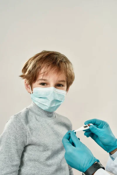Küçük çocuk doktoru ziyaret ediyor. Koruyucu maskeli küçük mutlu çocuk kameraya bakarken doktor da steril eldiven giyip sıcaklığı ölçüyor. — Stok fotoğraf