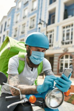 Kaliteli hizmet. Covid19 virüsünün ortaya çıkmasından dolayı maske ve eldiven takan teslimatçı motosiklet sürerken akıllı telefon kullanarak siparişleri kontrol ediyor. Kurye, dağıtım servisi, tecrit konsepti