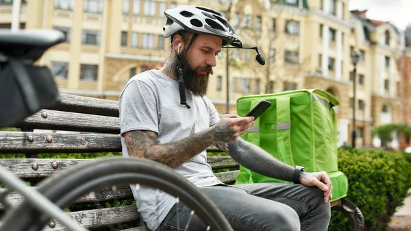 Zrób to po swojemu. Brutalny brodaty dostawca w kasku z torbą termiczną lub plecakiem sprawdzający kolejność za pomocą smartfona, siedzący na ławce na zewnątrz. Kurier, koncepcja usługi dostawy — Zdjęcie stockowe