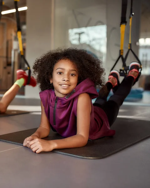 Tini lány nézi a kamerát, miközben gyakorolja a matracon segítségével fitness szíjak tornaterem — Stock Fotó