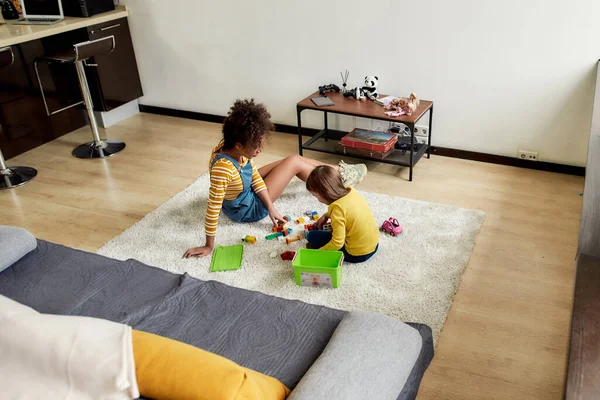 Menina caucasiana passando tempo com babá afro-americano. Eles estão brincando com brinquedos de construção, sentados no chão — Fotografia de Stock