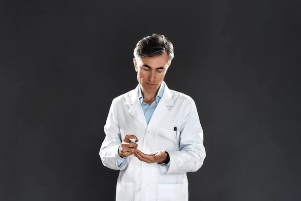 Szakmai érett férfi orvos orvosi egyenruhát használ fertőtlenítő gél vagy fertőtlenítő, miközben áll ellen fekete háttér — Stock Fotó