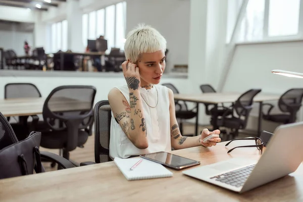 Rozmowa wideo. Młoda, stylowa blondynka wytatuowana kobieta biznesu zakładająca słuchawki bezprzewodowe siedząc w swoim miejscu pracy w nowoczesnym biurze — Zdjęcie stockowe