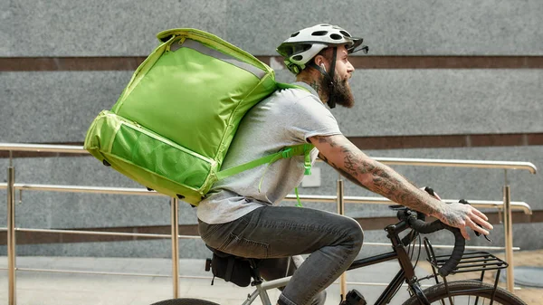 Food at anytime. Bearded delivery man in helmet with full thermo bag or backpack riding a bike along the city, delivering food. Courier, delivery service concept