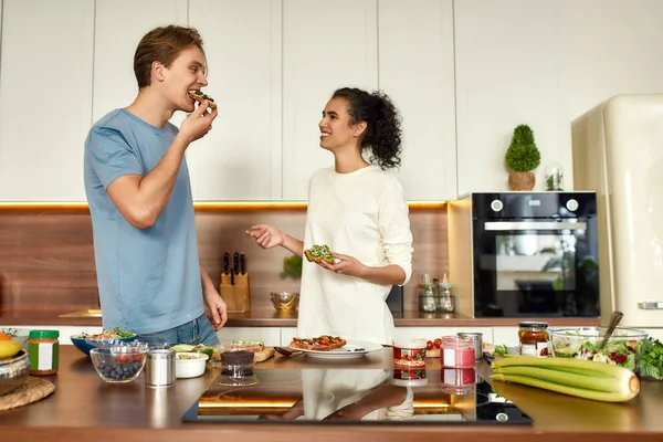 Είμαστε ό, τι τρώμε. Ευτυχισμένο ζευγάρι, χορτοφάγοι μαγειρεύουν πρωινό στην κουζίνα. Νεαρός άντρας και γυναίκα δοκιμάζουν σάντουιτς που έφτιαξαν μαζί. Χορτοφαγία, υγιεινά τρόφιμα, διατροφή, μείνετε στο σπίτι έννοια — Φωτογραφία Αρχείου