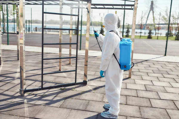 Providing cleanliness. Sanitization, cleaning and disinfection of the city due to the emergence of the Covid19 virus. Man in protective suit and mask at work near sports ground — Stock Photo, Image