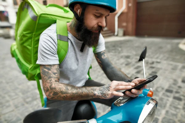 Το φαγητό σου είναι καθ 'οδόν. Γενειοφόρος διανομέας στέκεται με θερμική τσάντα ή σακίδιο σε εξωτερικούς χώρους, τον έλεγχο της παραγγελίας χρησιμοποιώντας smartphone, έτοιμος να οδηγήσει ένα σκούτερ κινητήρα — Φωτογραφία Αρχείου