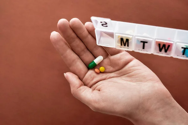 A weekly medicine dispenser opened for Sunday, hand holding pills and vitamins from white pill box on terracotta background. Health care, vitamins and treatment concept — Stock Photo, Image