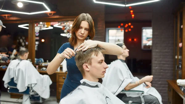 Barber meisje op het werk. Knappe jongeman in een kappersstoel terwijl het kappersmeisje voor hem knipt. Selectieve focus — Stockfoto