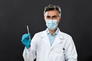 Üniformalı ve tıbbi maskeli olgun doktor ya da bilim adamı elinde grip aşısı, koronavirüs, covid-19 şırıngası tutarken siyah arka planda duruyor.