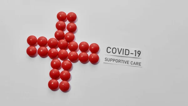 Top view of cross made of red pills and inscription Covid 19 Supportive Care lying diagonally on light background. Medications, virus and treatment concept — Stock Photo, Image