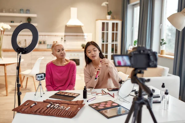 Ώρα να γίνουμε κοινωνικοί. Ασιάτισσα blogger που δείχνει προϊόντα ενώ βάζει μακιγιάζ σε μεσήλικες γυναίκες χρησιμοποιώντας καλλυντικά στο τραπέζι. Δύο γυναίκες καταγράφουν ένα εκπαιδευτικό βίντεο για το blog ομορφιάς — Φωτογραφία Αρχείου