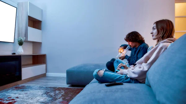 Vista lateral de un joven caucásico encendiendo cannabis en el bol de la pipa de agua de vidrio o pipa mientras juega videojuegos con un amigo, que se sientan en el sofá en casa —  Fotos de Stock