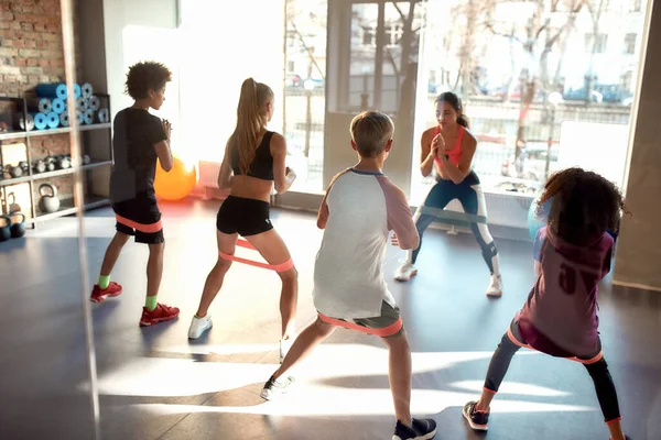 I bambini che si allenano con la fascia di resistenza in palestra insieme all'allenatore donna. Sport, stile di vita sano, educazione fisica — Foto Stock