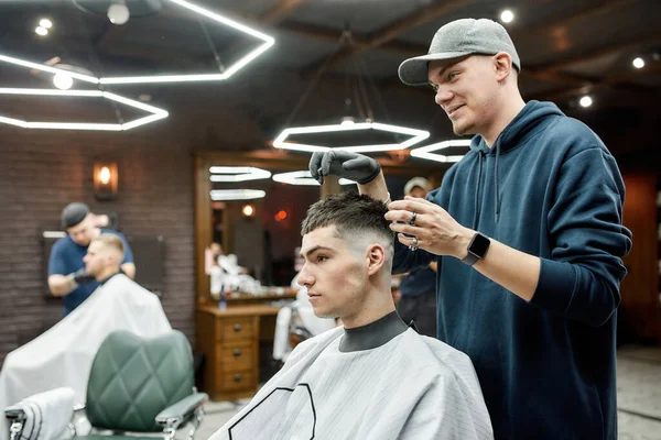 Lavoro in un barbiere. Barbiere professionista con un berretto da baseball che crea acconciature per un ragazzo giovane e bello — Foto Stock