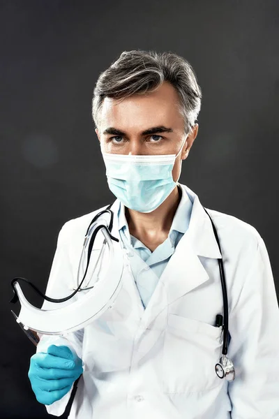 코로나 바이러스를 막아. 의료 마스크를 착용 한 성숙 한 남성 의사와 안면 보호 장갑을 끼고 어두운 배경에 맞서서 카메라를 보고 있는 모습. 사람에 초점을 맞추라 — 스톡 사진