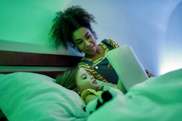 Una infancia alegre dura para siempre. Mujer afroamericana niñera y linda niña leyendo libro en la noche. Niño sosteniendo libro y mirando fotos —  Fotos de Stock