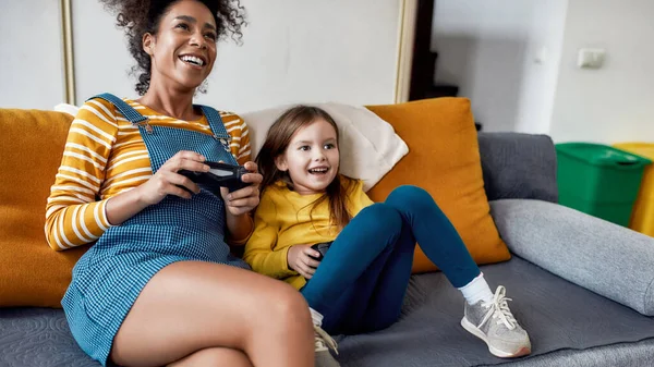 Pengurus terbaik. Wanita Afrika Amerika, pengasuh bayi dan gadis kecil bersenang-senang bersama, bermain video game, duduk di sofa. Pendidikan anak, kegiatan rekreasi, konsep mengasuh anak — Stok Foto