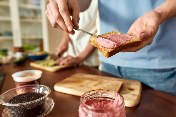 Close up de homem espalhando hummus beterraba rosa sobre pão torrado. Casal de vegetarianos preparando refeição saudável na cozinha juntos. Vegetarianismo, conceito de alimentação saudável — Fotografia de Stock