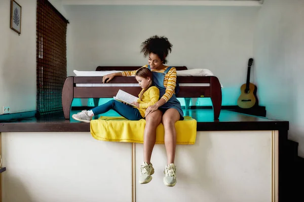 Подружитесь с книгой. Африканская американская няня и кавказская девочка читают книгу, сказку вместе сидя дома. Образование для детей, досуг, концепция няни — стоковое фото