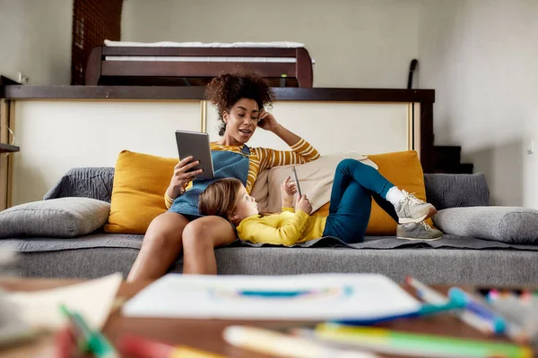 Hämta mer. Afrikansk amerikansk kvinna barnvakt och kaukasisk söt liten flicka med hjälp av surfplatta dator, sitter på soffan. Utbildning för barn, fritidsaktiviteter, barnpassning — Stockfoto