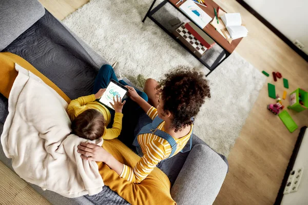 Het realiseren van fantasie. Afro-Amerikaanse vrouw babysitter vermaken Kaukasisch schattig klein meisje. Kind tekent met tablet pc, zittend op de bank — Stockfoto