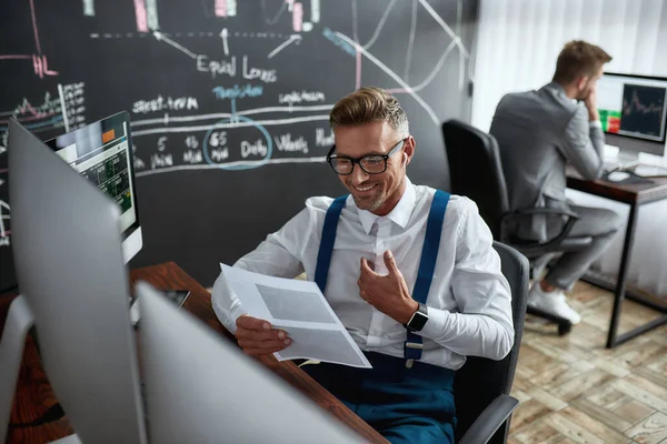 Taak ontwerpen voor geld. Stijlvolle zakenman, handelaar die papieren doorneemt, terwijl hij achter een bureau zit voor een computermonitor. Zijn collega werkt op de achtergrond — Stockfoto