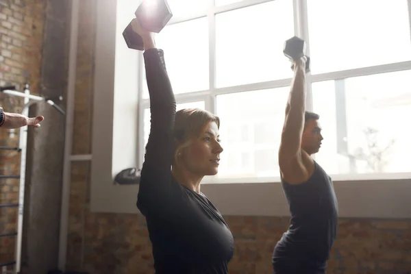 Prepárate para estar en forma. Mujer atlética levantando mancuerna mientras hace ejercicio en el gimnasio industrial. Formación en grupo, concepto de trabajo en equipo —  Fotos de Stock