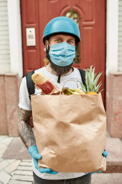 Bearded delivery man in helmet wearing mask and gloves due to the emergence of the Covid19 virus, holding grocery bag, while delivering food. Courier, delivery service, lockdown concept Royalty Free Stock Photos